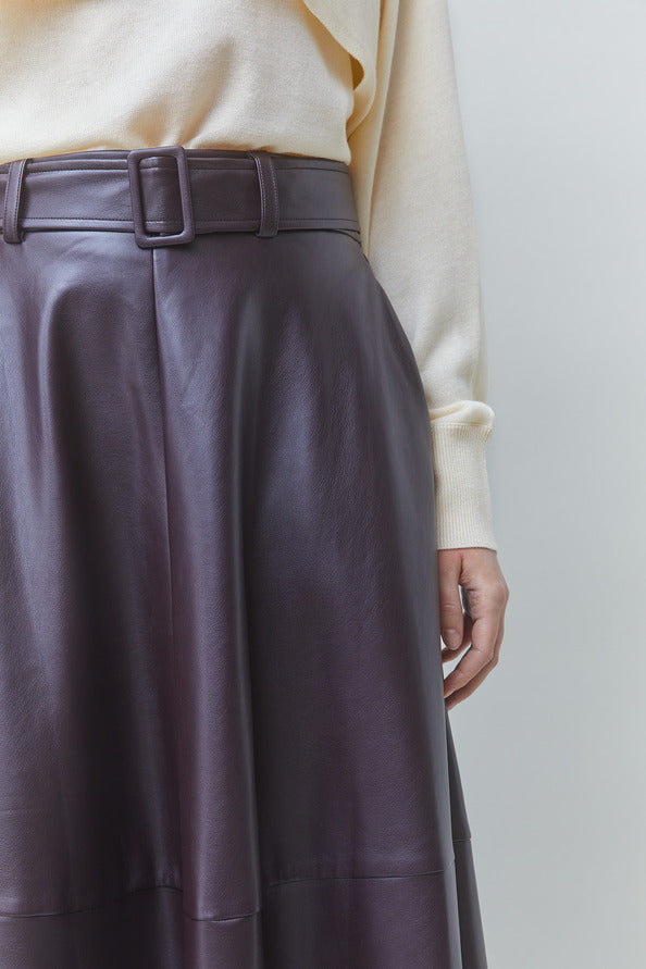 Close-up of a person wearing a dark plum faux-leather skirt with a matching belt and a cream ribbed sweater, against a light gray background.