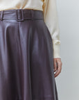 Close-up of a person wearing a dark plum faux-leather skirt with a matching belt and a cream ribbed sweater, against a light gray background.