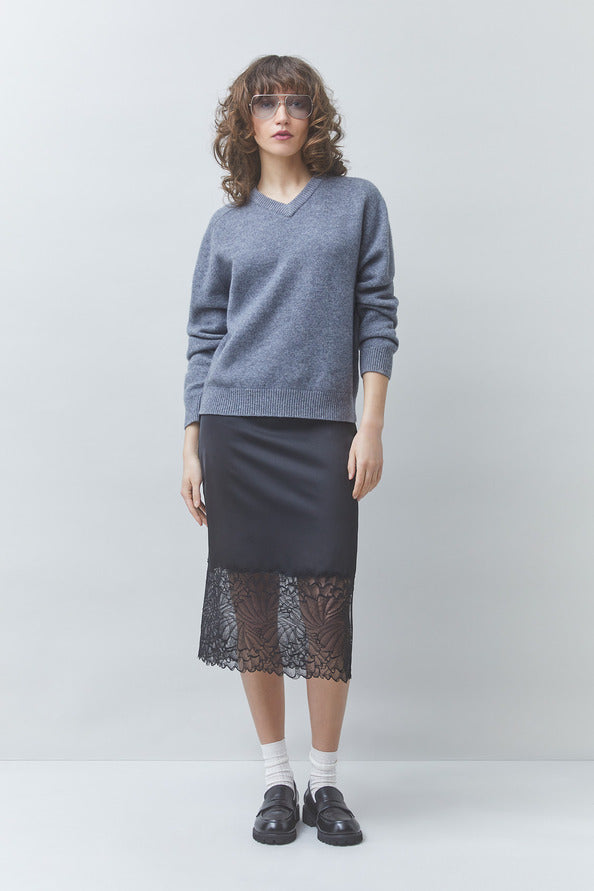 A model stands against a light gray studio background wearing a heathered blue-gray V-neck sweater, a dark charcoal skirt with a black lace hem, white socks, and black loafers.