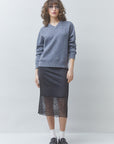 A model stands against a light gray studio background wearing a heathered blue-gray V-neck sweater, a dark charcoal skirt with a black lace hem, white socks, and black loafers.