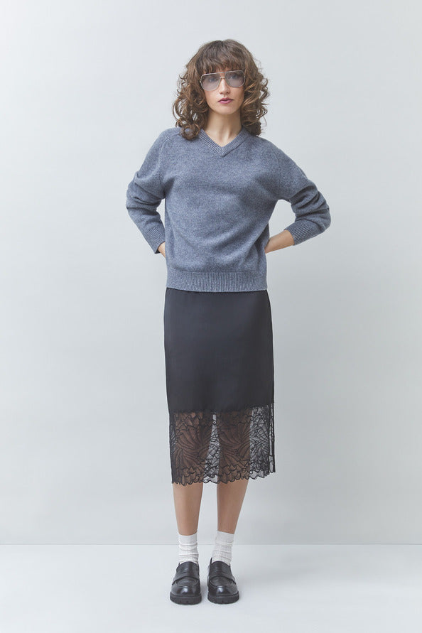 A model stands against a light gray studio background wearing a heathered blue-gray V-neck sweater, a black midi skirt with a sheer black lace hem, white socks, and black loafers.