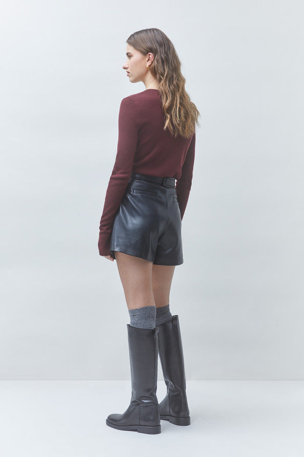A woman is shown from the back wearing a burgundy long-sleeve top, black leather shorts, gray socks, and black knee-high boots against a light gray studio background.