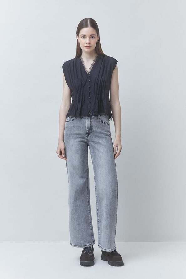 A woman stands against a light gray studio background wearing a sleeveless dark pleated top with front buttons, light gray wide-leg jeans, and dark shoes.