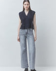 A woman stands against a light gray studio background wearing a sleeveless dark pleated top with front buttons, light gray wide-leg jeans, and dark shoes.