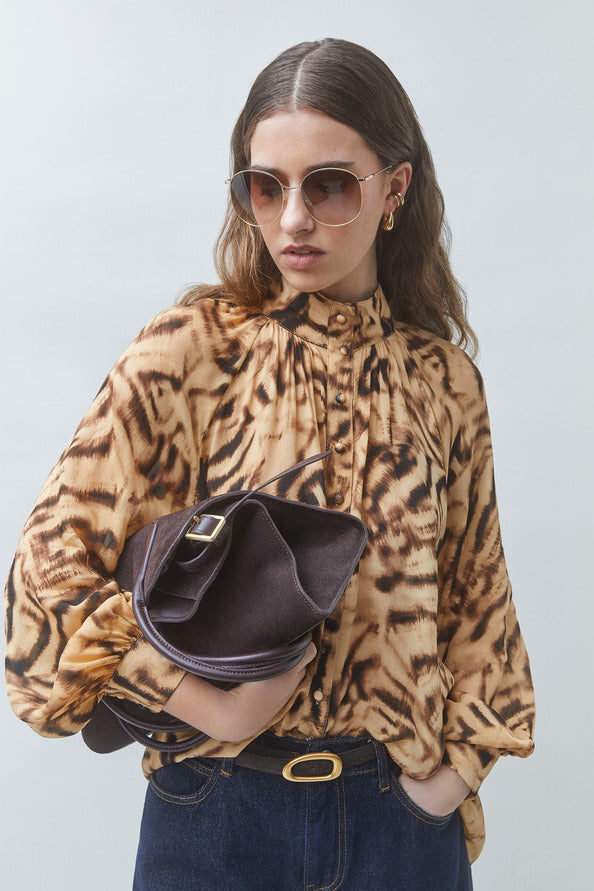 A woman wearing a brown and beige animal-print blouse, dark sunglasses, and a dark brown handbag, standing against a light gray studio background.