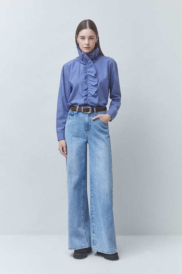 A model stands against a light gray studio background wearing a blue and white gingham long-sleeve blouse with a high ruffled mock neck, paired with wide-leg light blue jeans and a dark belt.