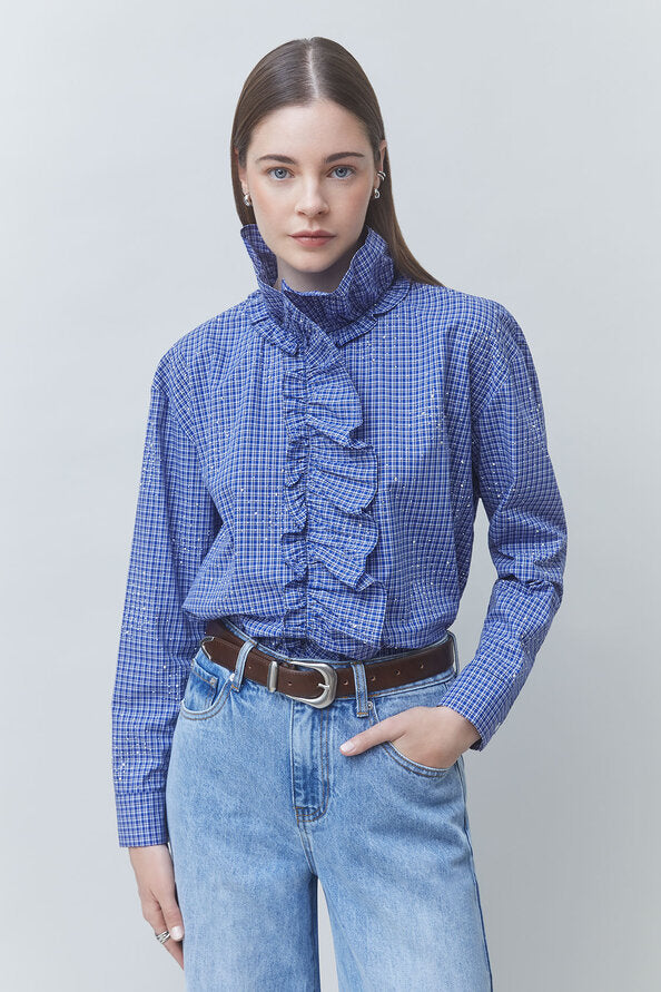 A woman modeling a blue and white gingham long-sleeve blouse with a high ruffled mock neck, paired with light-wash blue jeans and a brown belt, against a solid light gray background.