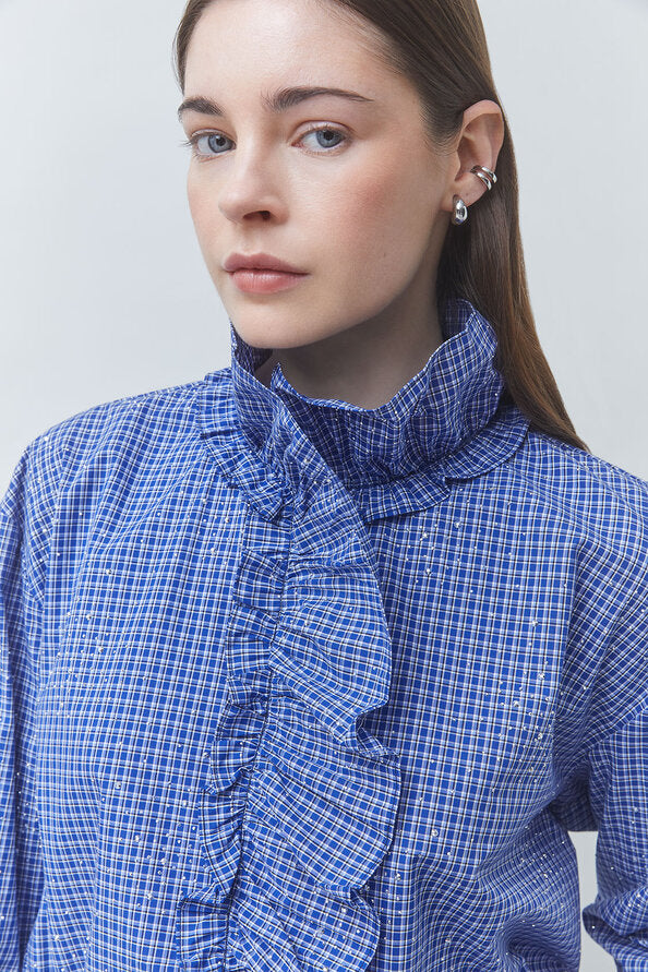 A close-up of a woman wearing a blue and white checkered blouse with a high ruffled mock neck collar. She is wearing silver hoop earrings against a plain light gray background.
