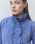 A close-up of a woman wearing a blue and white checkered blouse with a high ruffled mock neck collar. She is wearing silver hoop earrings against a plain light gray background.