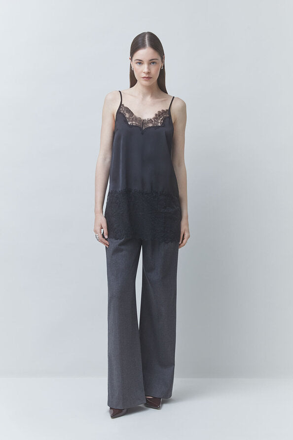A woman stands against a light gray studio background wearing a dark gray sleeveless jumpsuit with black lace trim at the neckline and hem, paired with dark pointed shoes.
