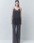 A woman stands against a light gray studio background wearing a dark gray sleeveless jumpsuit with black lace trim at the neckline and hem, paired with dark pointed shoes.