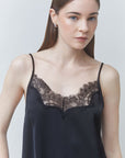 A woman wearing a black satin camisole with a black lace V-neckline, photographed against a light gray studio background.