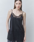 A woman modeling a black satin camisole top with black lace trim at the neckline and hem, paired with gray pants, against a light gray studio background.
