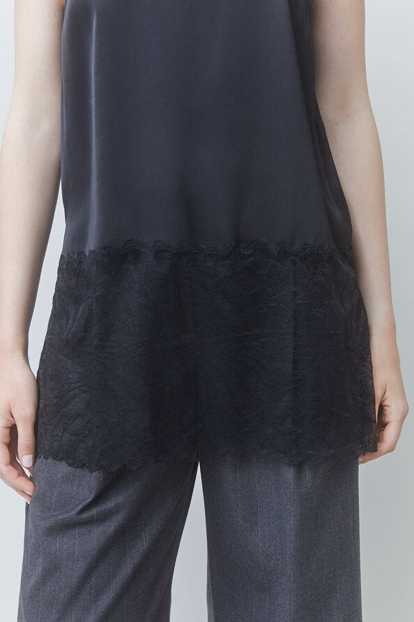 A close-up of a person wearing a black sleeveless top with a sheer mesh upper section and a textured lace ruffle hem, paired with gray trousers.