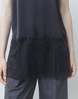 A close-up of a person wearing a black sleeveless top with a sheer mesh upper section and a textured lace ruffle hem, paired with gray trousers.