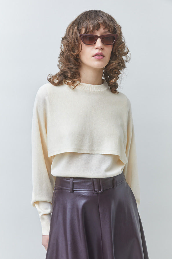 A model with curly brown hair and sunglasses wearing a cream-colored cropped long-sleeve knit top and a dark purple high-waisted leather skirt, set against a plain light gray studio background.