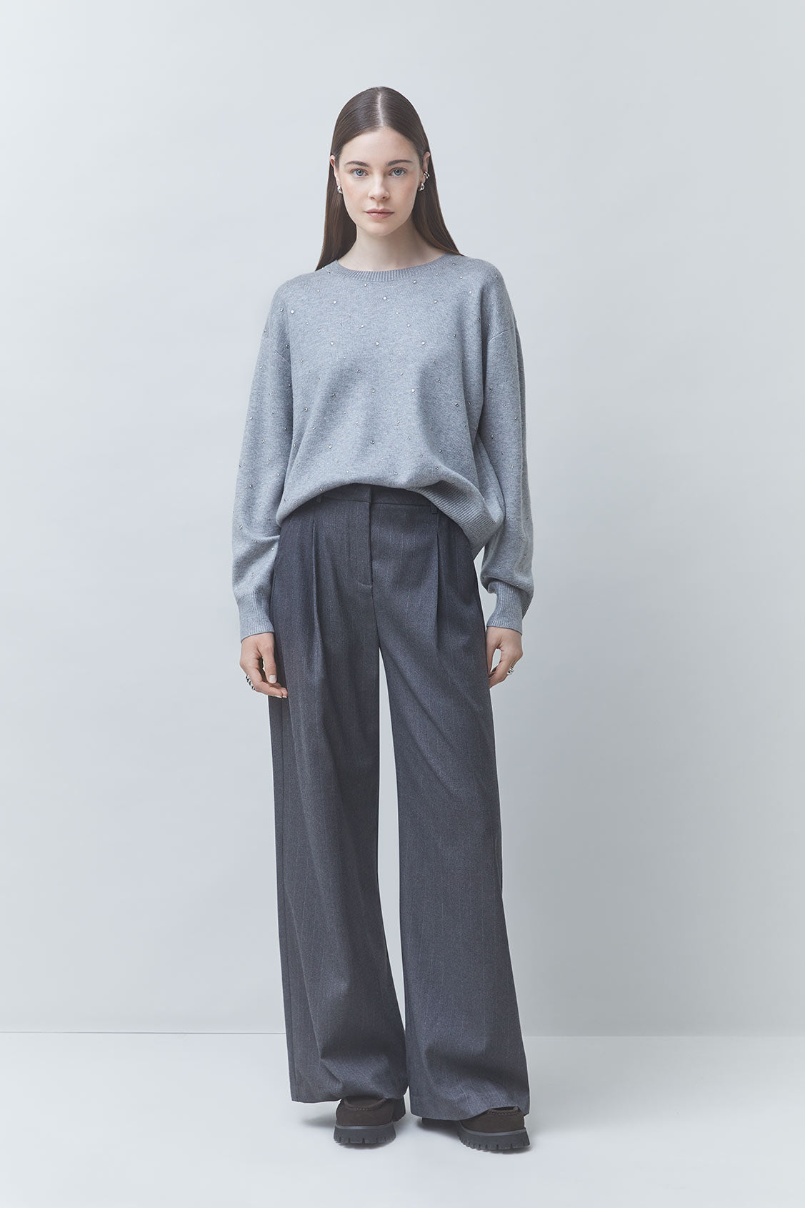 A woman stands against a light gray studio background wearing a light gray knit sweater and dark gray wide-leg trousers.