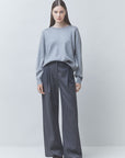 A woman stands against a light gray studio background wearing a light gray knit sweater and dark gray wide-leg trousers.
