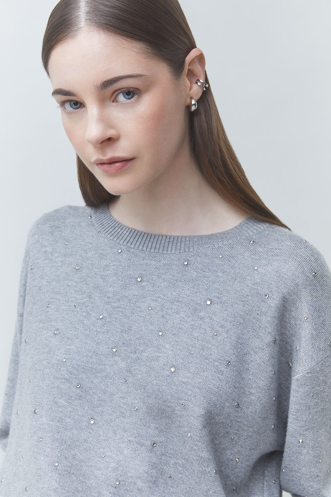 A woman wearing a light gray knit sweater adorned with small silver rhinestones, photographed against a plain light gray background.