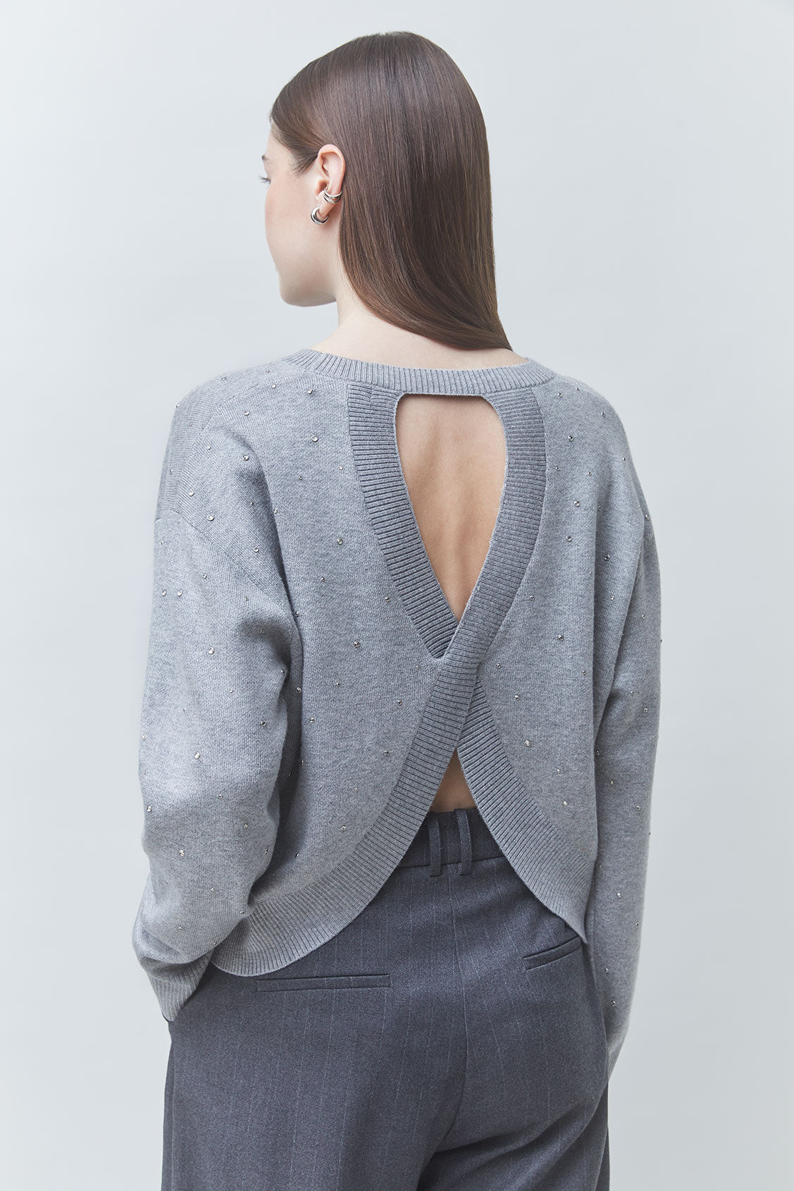 A woman shown from the back wearing a light gray knit sweater with a large open V-back design and dark gray trousers, photographed against a plain light gray studio background.