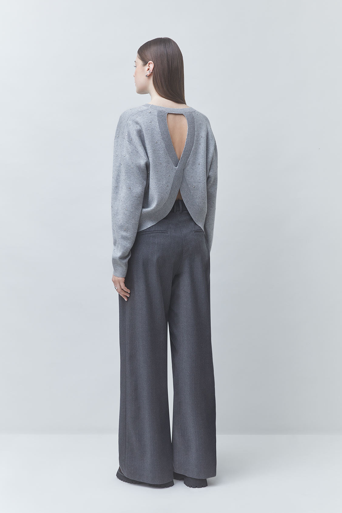 A model shown from the back wearing a light gray knit long-sleeve top with a crossover open back and dark gray wide-leg trousers, set against a plain light gray studio background.