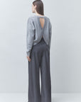 A model shown from the back wearing a light gray knit long-sleeve top with a crossover open back and dark gray wide-leg trousers, set against a plain light gray studio background.