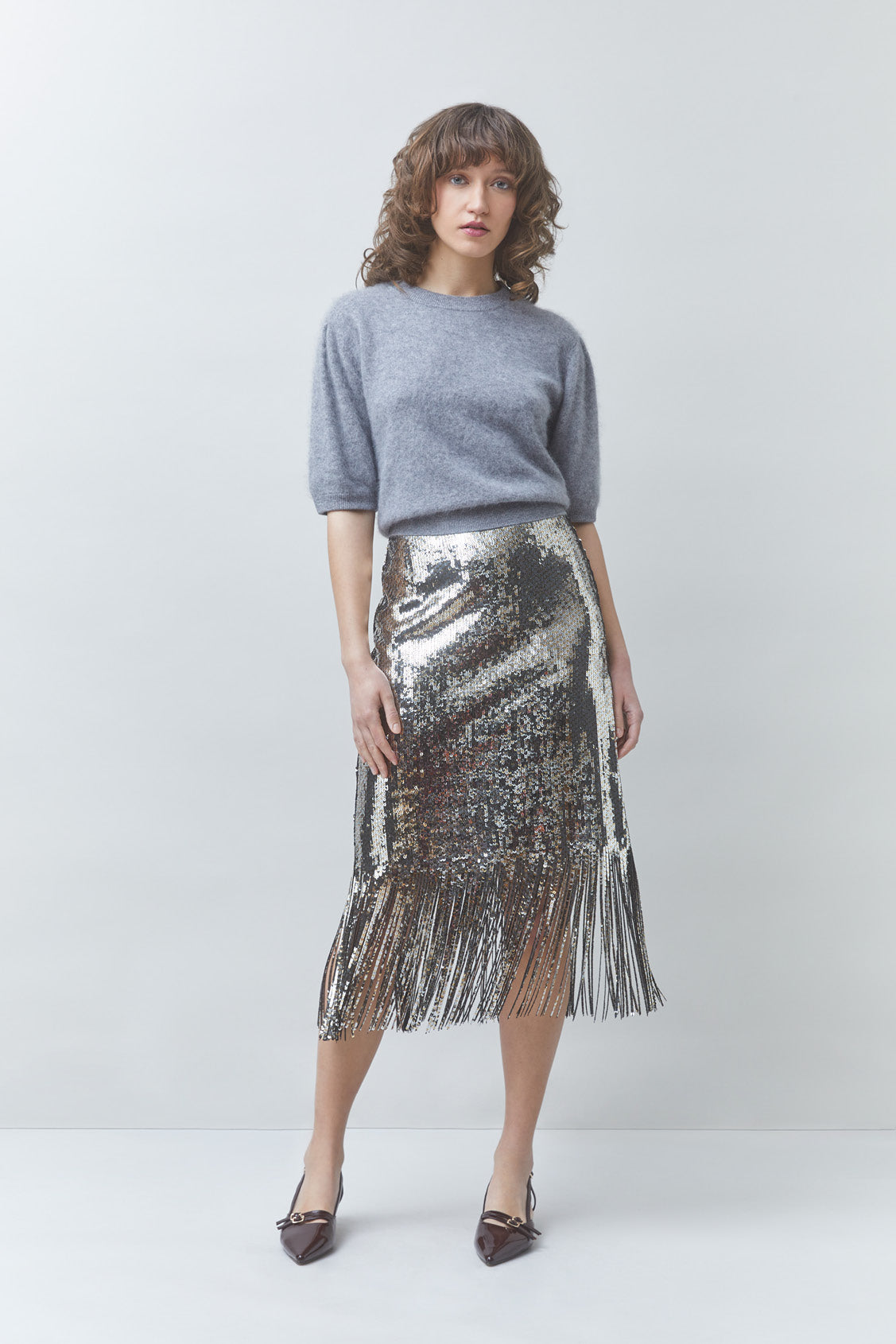 A woman modeling a gray knit short-sleeve top and a metallic silver sequined midi skirt with fringe, paired with dark pointed shoes, against a light gray studio background.