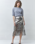 A woman modeling a gray knit short-sleeve top and a metallic silver sequined midi skirt with fringe, paired with dark pointed shoes, against a light gray studio background.