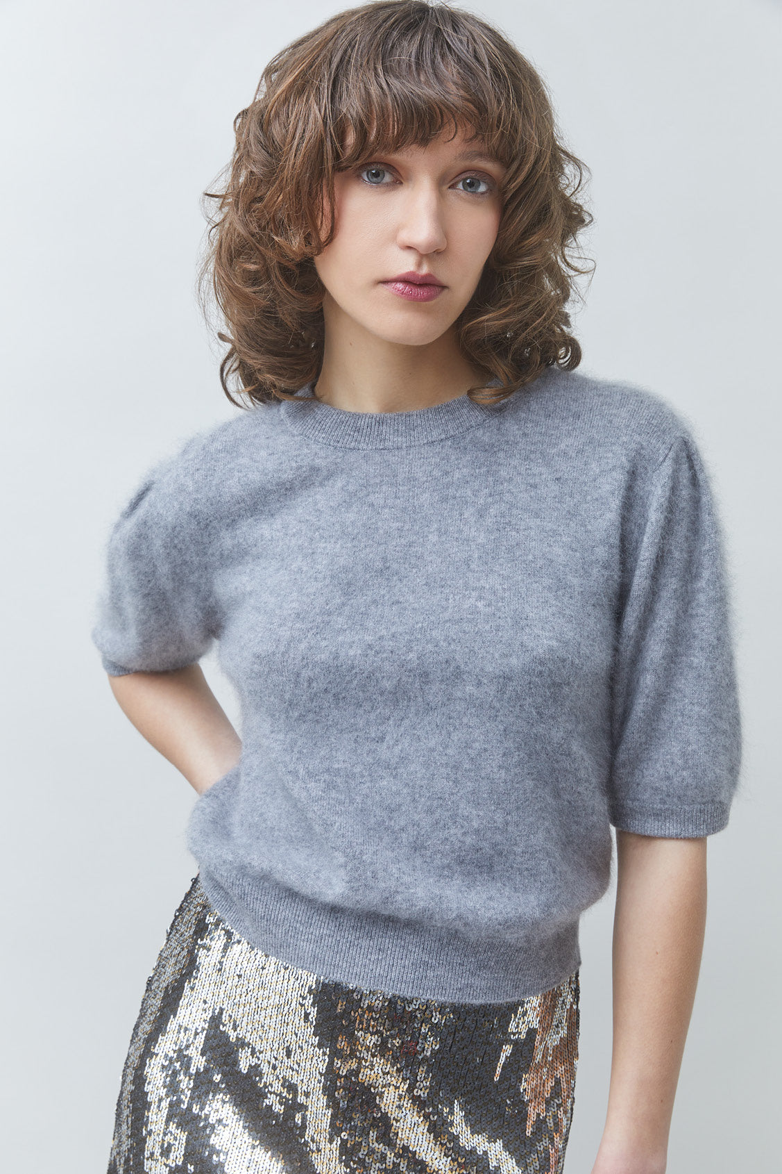 A woman with shoulder-length brown hair wearing a light gray short-sleeve knit sweater and a patterned metallic skirt, photographed against a plain light gray studio background.