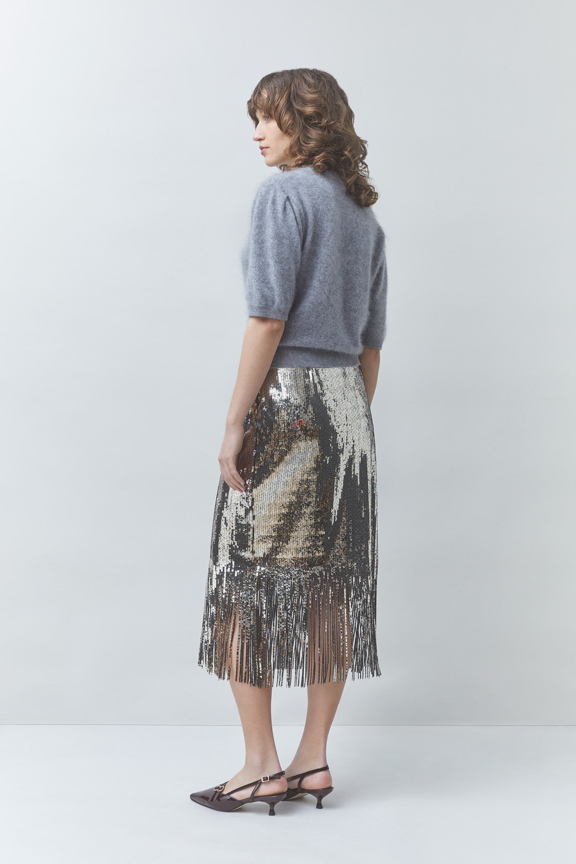A woman stands in profile wearing a gray short-sleeve knit top and a metallic silver sequined midi skirt with fringe, paired with dark heeled sandals, against a light gray studio background.