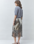 A woman stands in profile wearing a gray short-sleeve knit top and a metallic silver sequined midi skirt with fringe, paired with dark heeled sandals, against a light gray studio background.