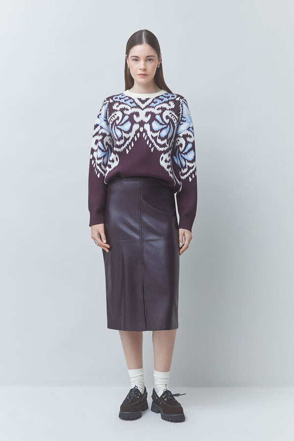 A model stands against a light gray studio background wearing a long-sleeve knit sweater with a dark purple base and ornate white and light blue patterned yoke, paired with a dark purple midi skirt, white socks, and black shoes.