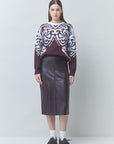 A model stands against a light gray studio background wearing a long-sleeve knit sweater with a dark purple base and ornate white and light blue patterned yoke, paired with a dark purple midi skirt, white socks, and black shoes.