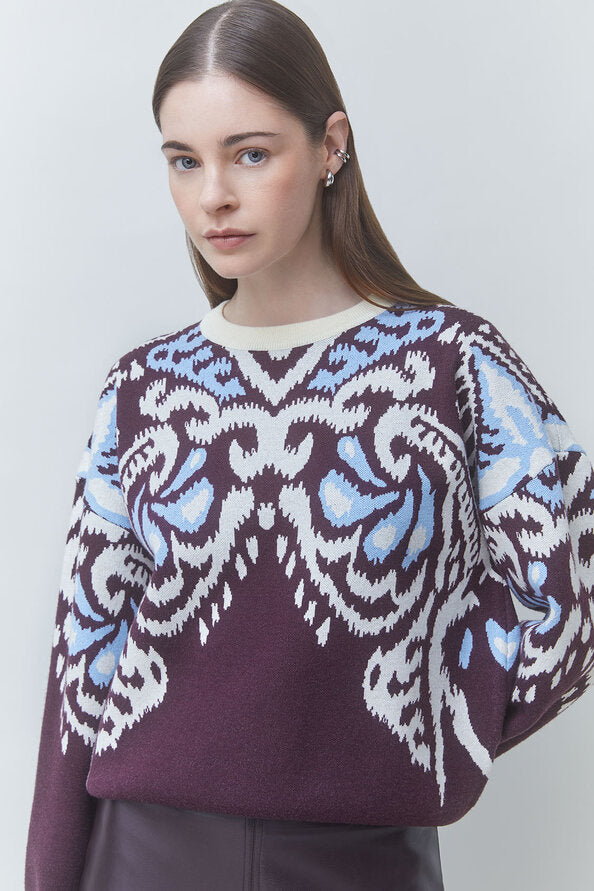 A woman wearing a dark purple knit sweater with a white and light blue ornate pattern across the chest and sleeves, paired against a light gray studio background.