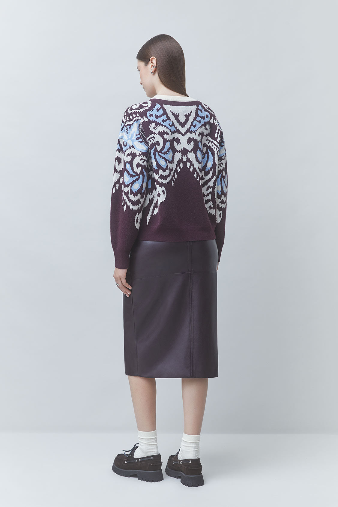A model is shown from the back wearing a dark purple knit sweater with a white and light blue ornate pattern, paired with a dark purple midi skirt, white socks, and black loafers, against a light gray studio background.