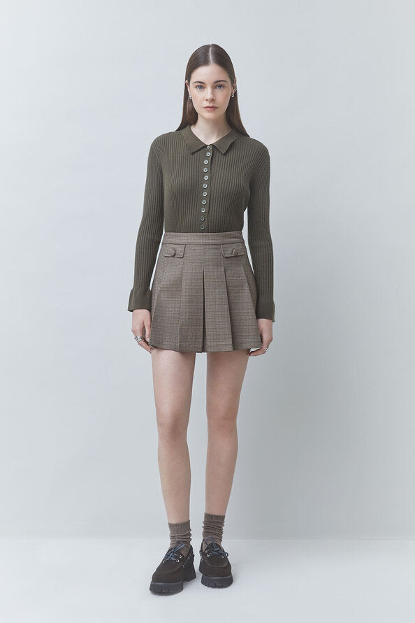 A model stands against a light gray studio background wearing a dark olive green ribbed long-sleeve polo shirt with a button placket and a brown-and-gray plaid pleated mini skirt, paired with gray socks and dark brown shoes.