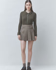 A model stands against a light gray studio background wearing a dark olive green ribbed long-sleeve polo shirt with a button placket and a brown-and-gray plaid pleated mini skirt, paired with gray socks and dark brown shoes.