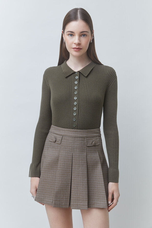 A woman modeling a ribbed long-sleeve polo-knit top in olive green paired with a brown plaid pleated mini skirt, shot against a light gray studio background.