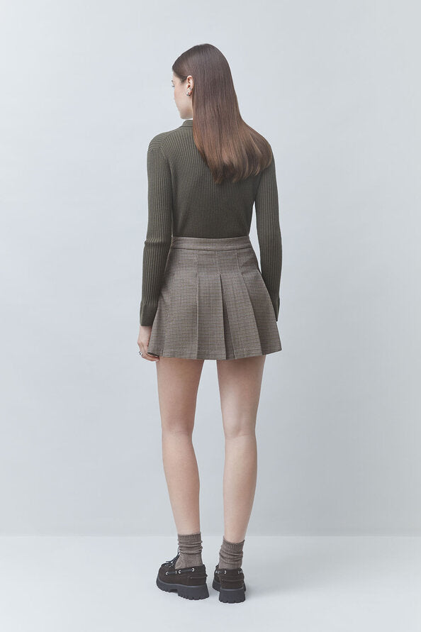 A model shown from the back wearing an olive green ribbed long-sleeve top and a brown and gray plaid pleated mini skirt, paired with gray socks and black platform shoes, against a light gray studio background.