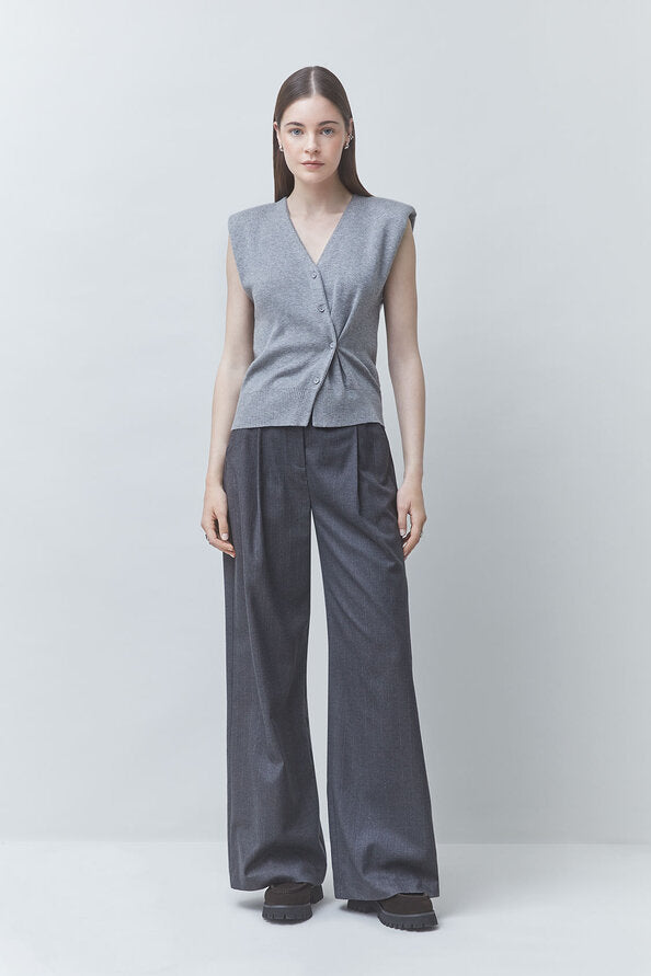 A model stands against a light gray studio background wearing a sleeveless wrap-style knit vest in heather gray with dark charcoal wide-leg trousers and black shoes.