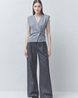 A model stands against a light gray studio background wearing a sleeveless wrap-style knit vest in heather gray with dark charcoal wide-leg trousers and black shoes.