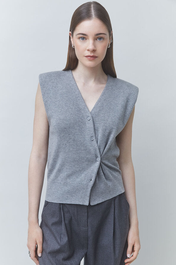 A woman modeling a sleeveless gray knit cardigan vest with a twisted front detail and button closure, paired with dark gray trousers, against a light gray studio background.