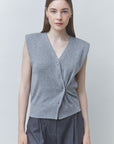 A woman modeling a sleeveless gray knit cardigan vest with a twisted front detail and button closure, paired with dark gray trousers, against a light gray studio background.