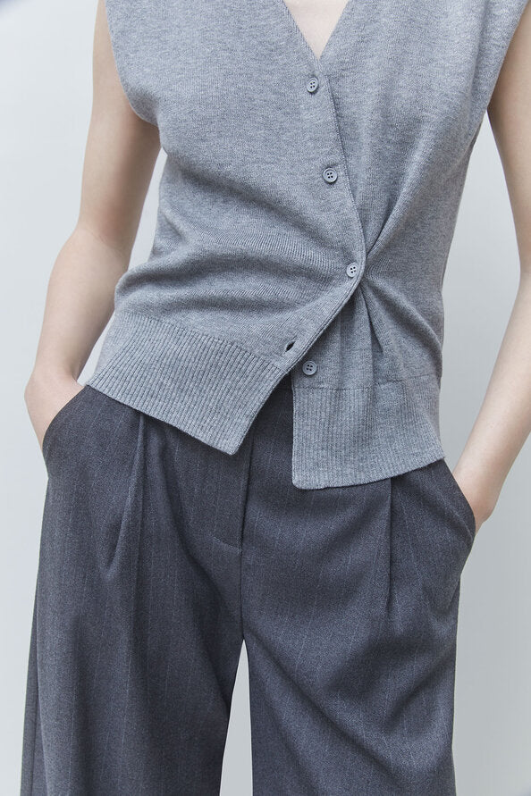 A close-up of a person wearing a sleeveless gray knit wrap vest with front buttons and dark gray wide-leg trousers, standing against a light gray studio background.