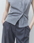 A close-up of a person wearing a sleeveless gray knit wrap vest with front buttons and dark gray wide-leg trousers, standing against a light gray studio background.