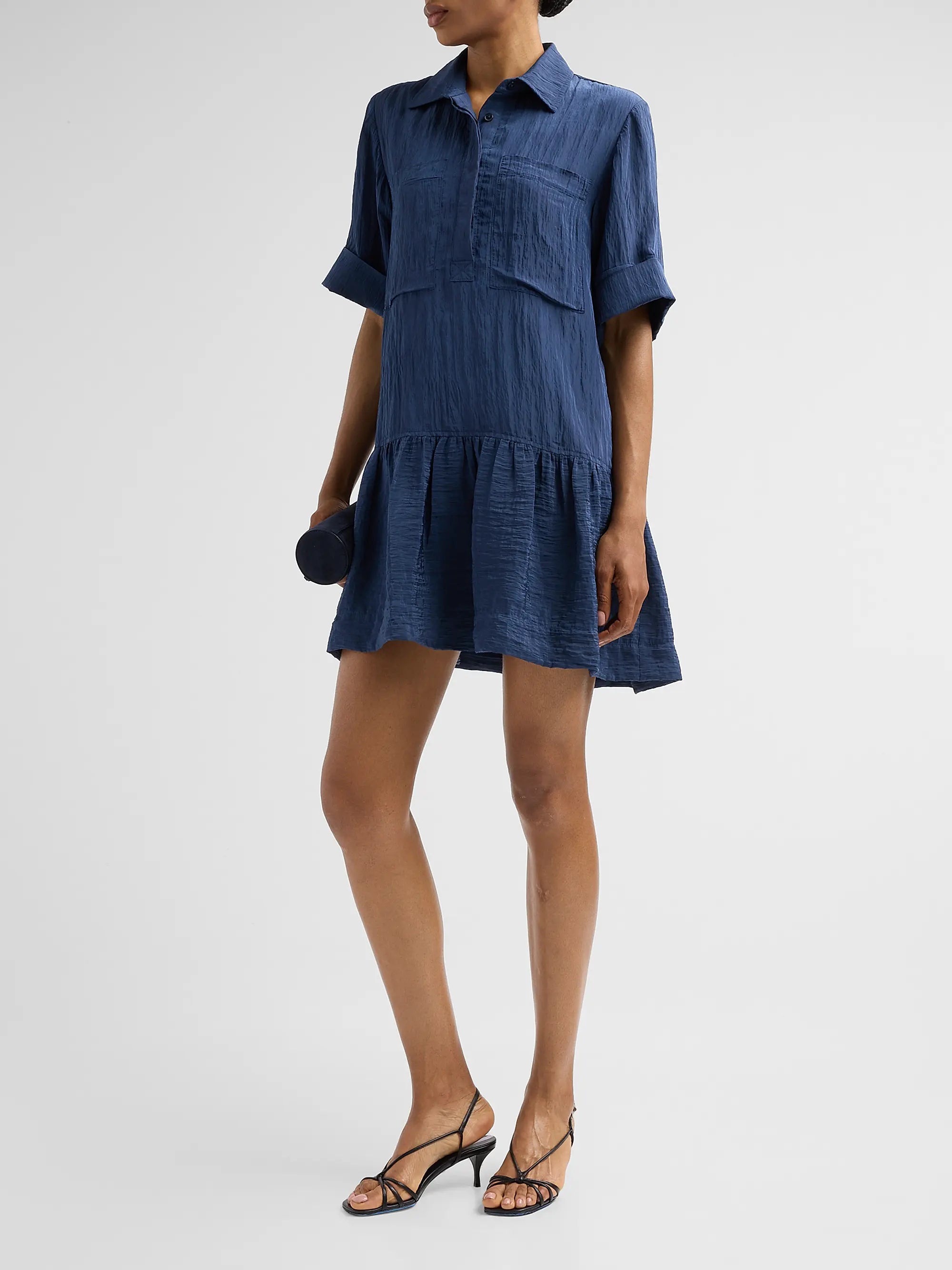 A model wearing a short-sleeve blue textured shirt dress with a button-up front and ruffled hem, paired with black strappy sandals against a light gray studio background.