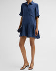 A model wearing a short-sleeve blue textured shirt dress with a button-up front and ruffled hem, paired with black strappy sandals against a light gray studio background.