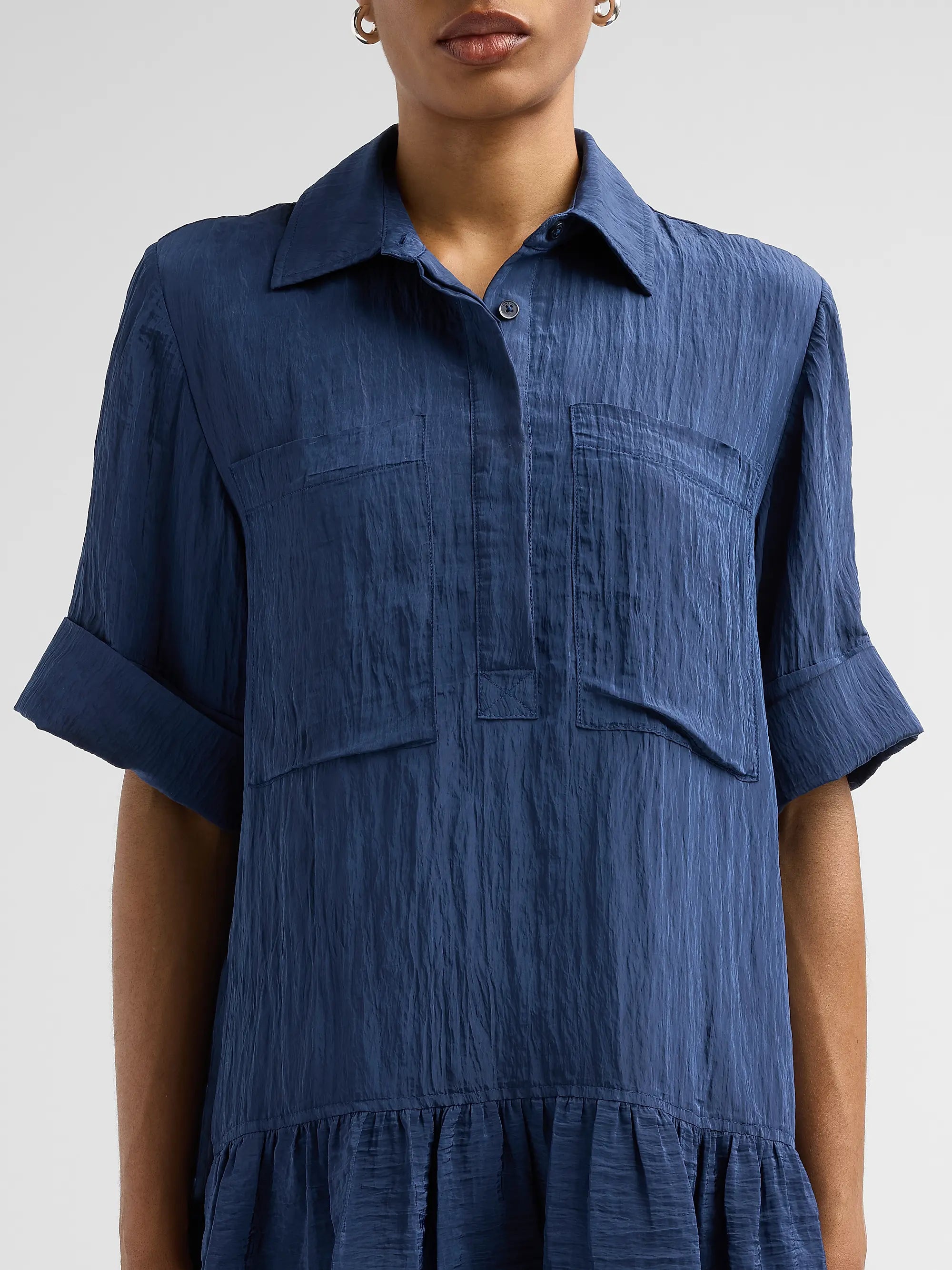 A woman wearing a textured blue short-sleeve collared shirt dress with rolled cuffs and a tiered hem, shown from the nose down against a light gray studio background.