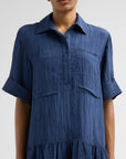 A woman wearing a textured blue short-sleeve collared shirt dress with rolled cuffs and a tiered hem, shown from the nose down against a light gray studio background.