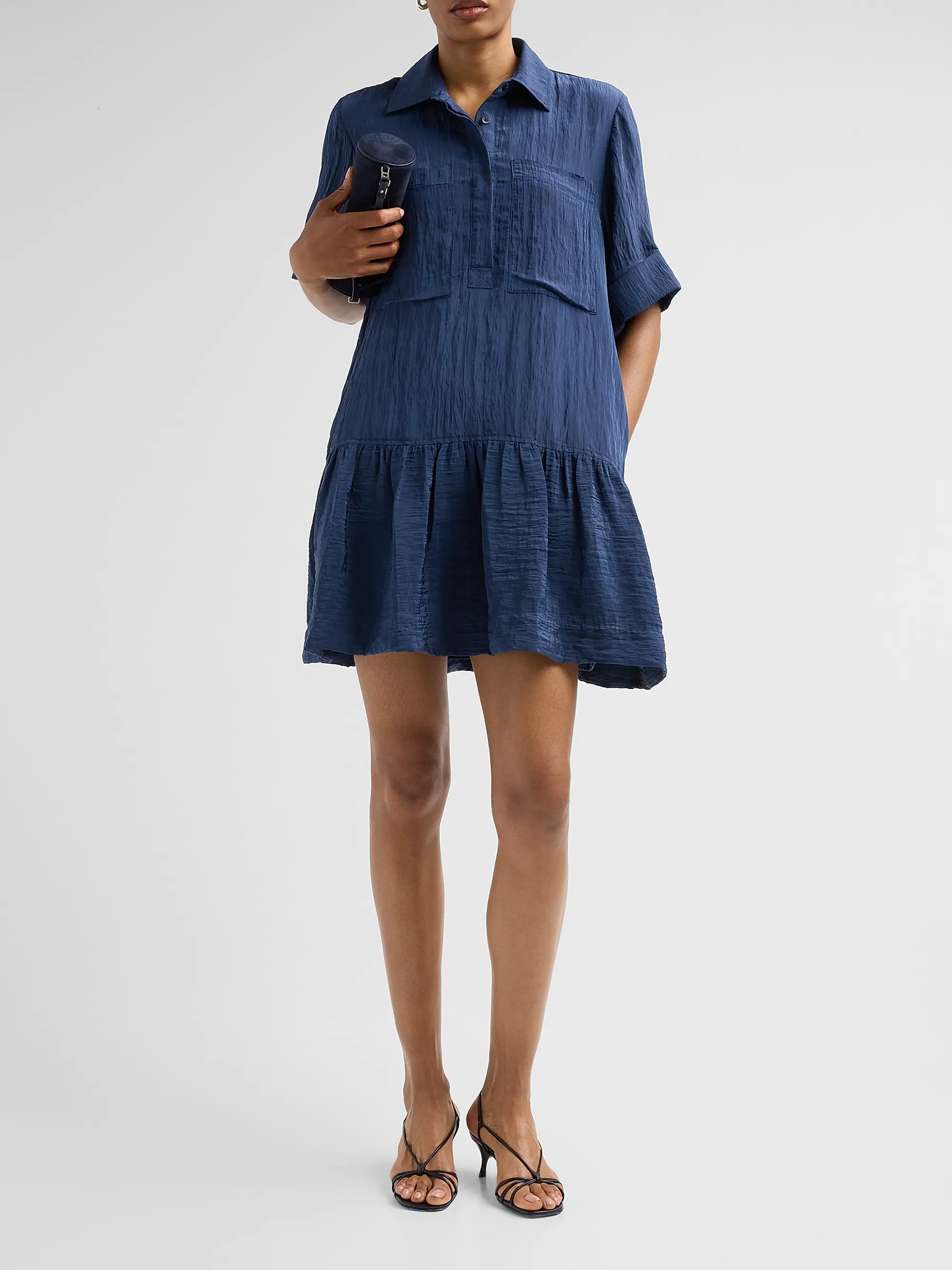 A woman modeling a short-sleeve blue tiered shirt dress with a textured crinkle fabric, paired with black strappy heels against a light gray studio background.